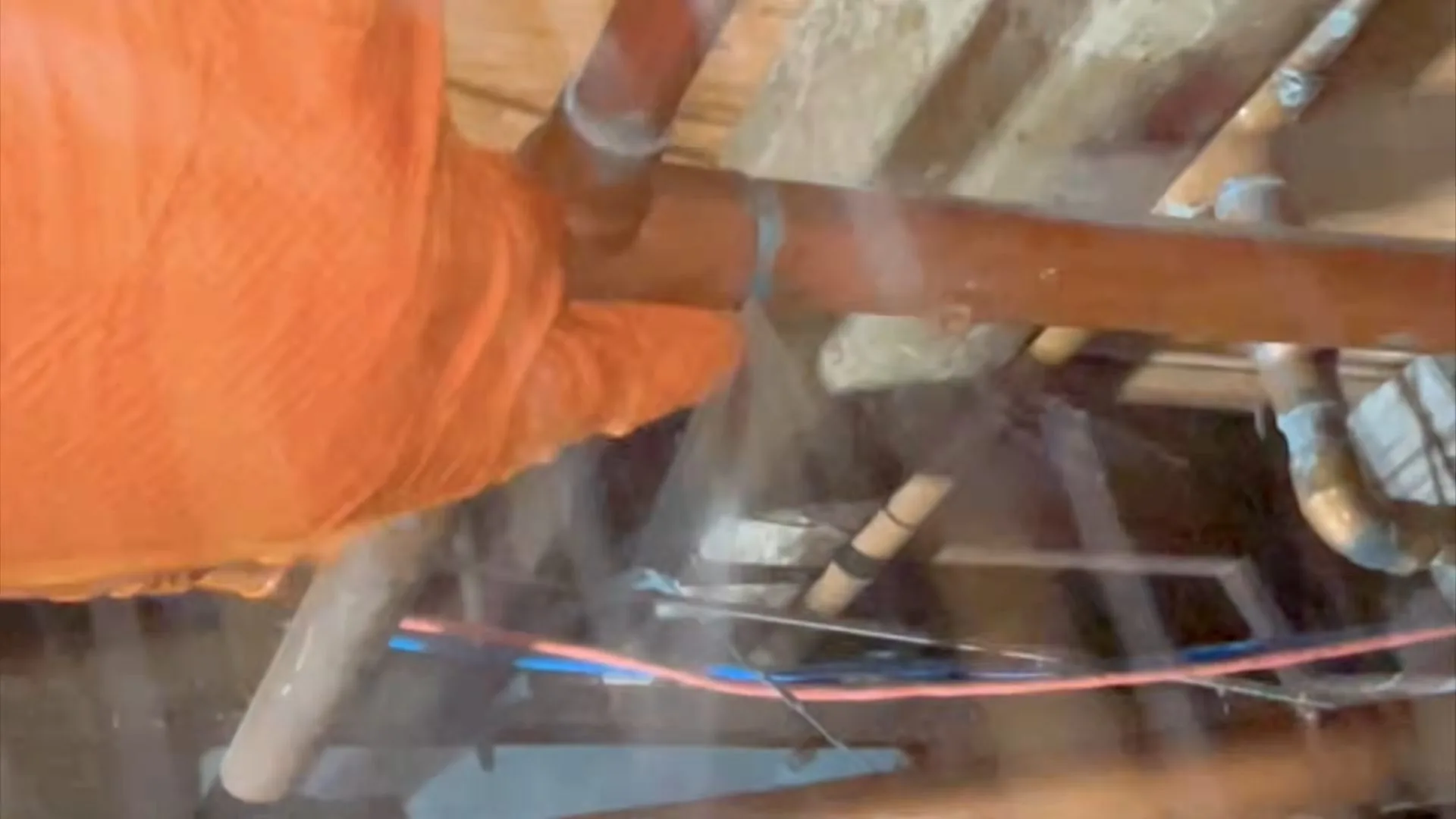 A gloved hand holding a copper supply pipe between floor joists with water spraying around the joint